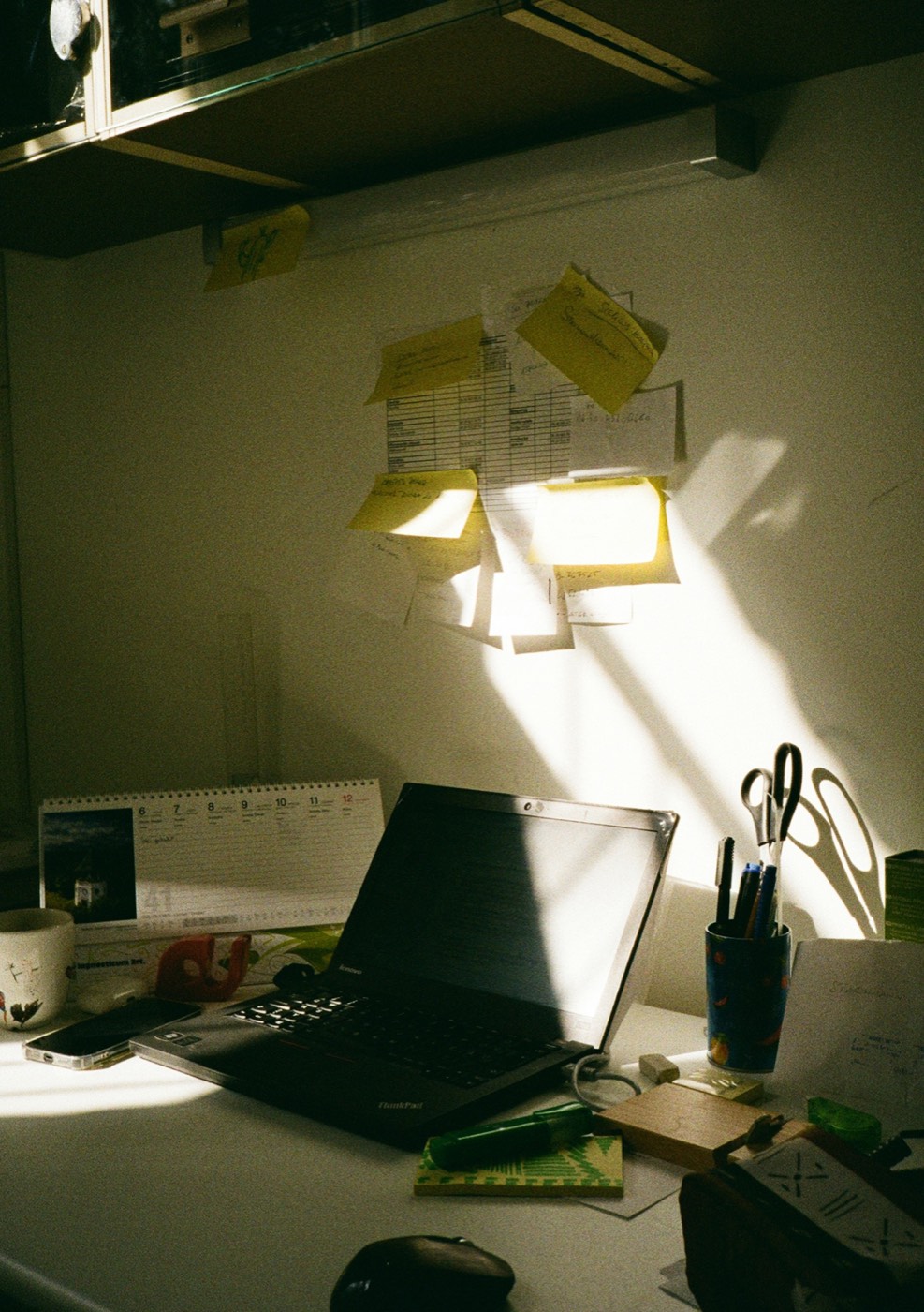 A small desk in afternoon sun: a laptop, a cup of pens, a desk calendar and sticky notes on the wall