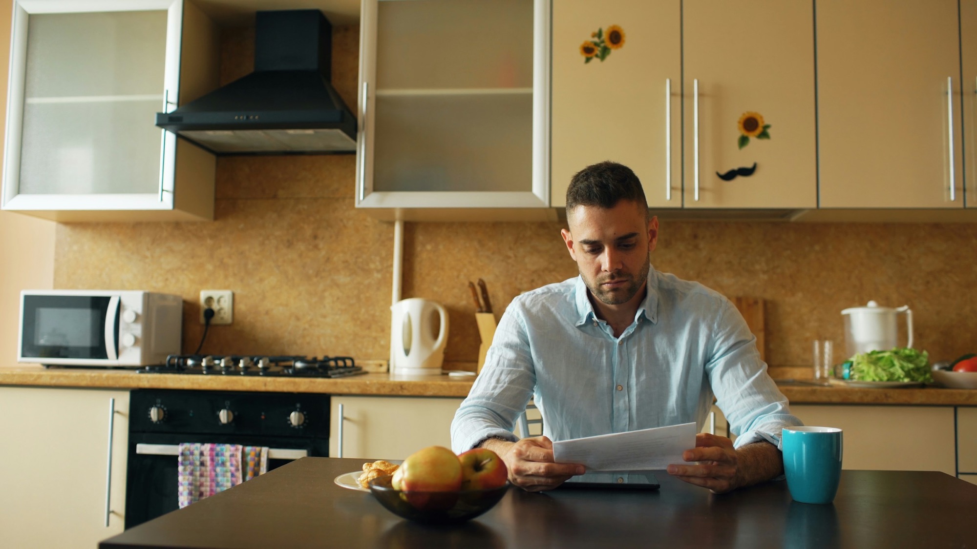 A man reading a printed page at his kitchen table in the morning, a mug and a bowl of fruit beside him