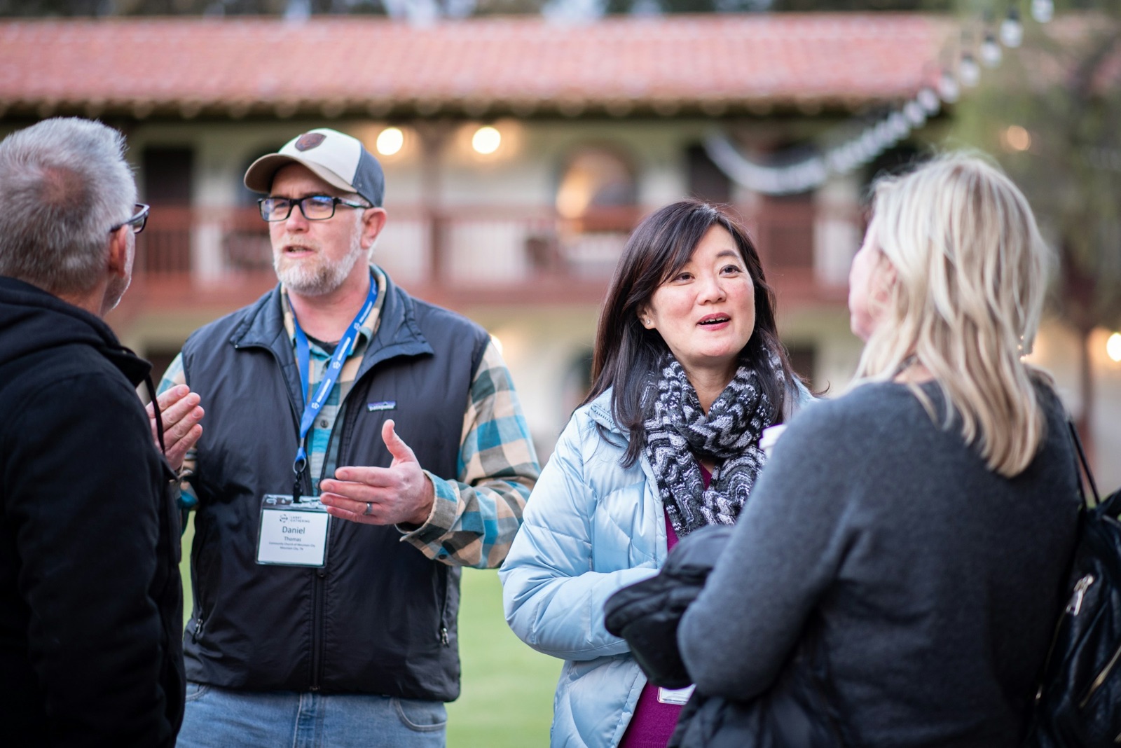 Three people with name badges in conversation outdoors at an event, a building with string lights behind them