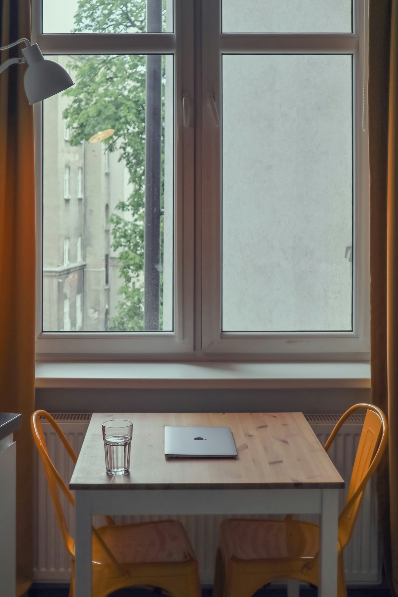 A small wooden table with a laptop and a glass of water in front of a tall window