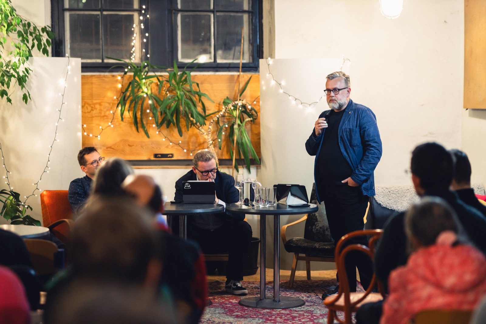 A host speaking to a small audience in a cozy room with plants and string lights, a live recording set up on the table
