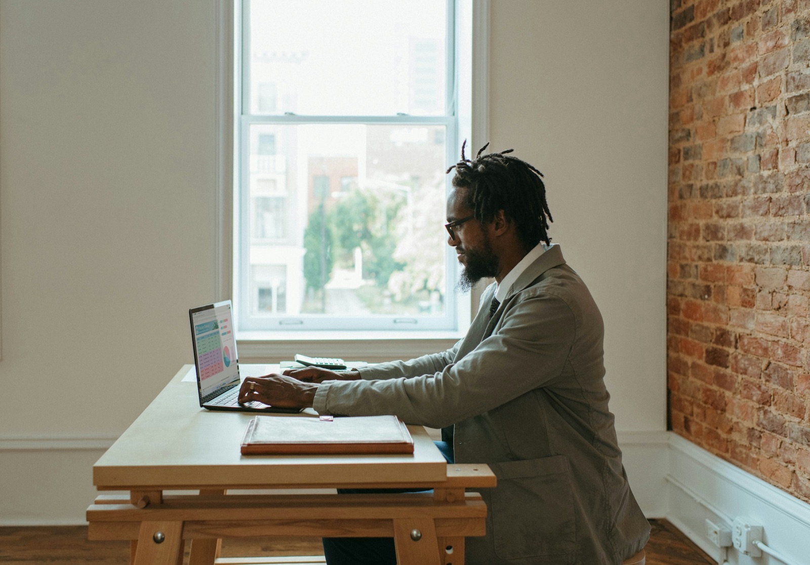 A man working on a laptop at a simple wooden desk beside a tall window and a brick wall, seen from the side