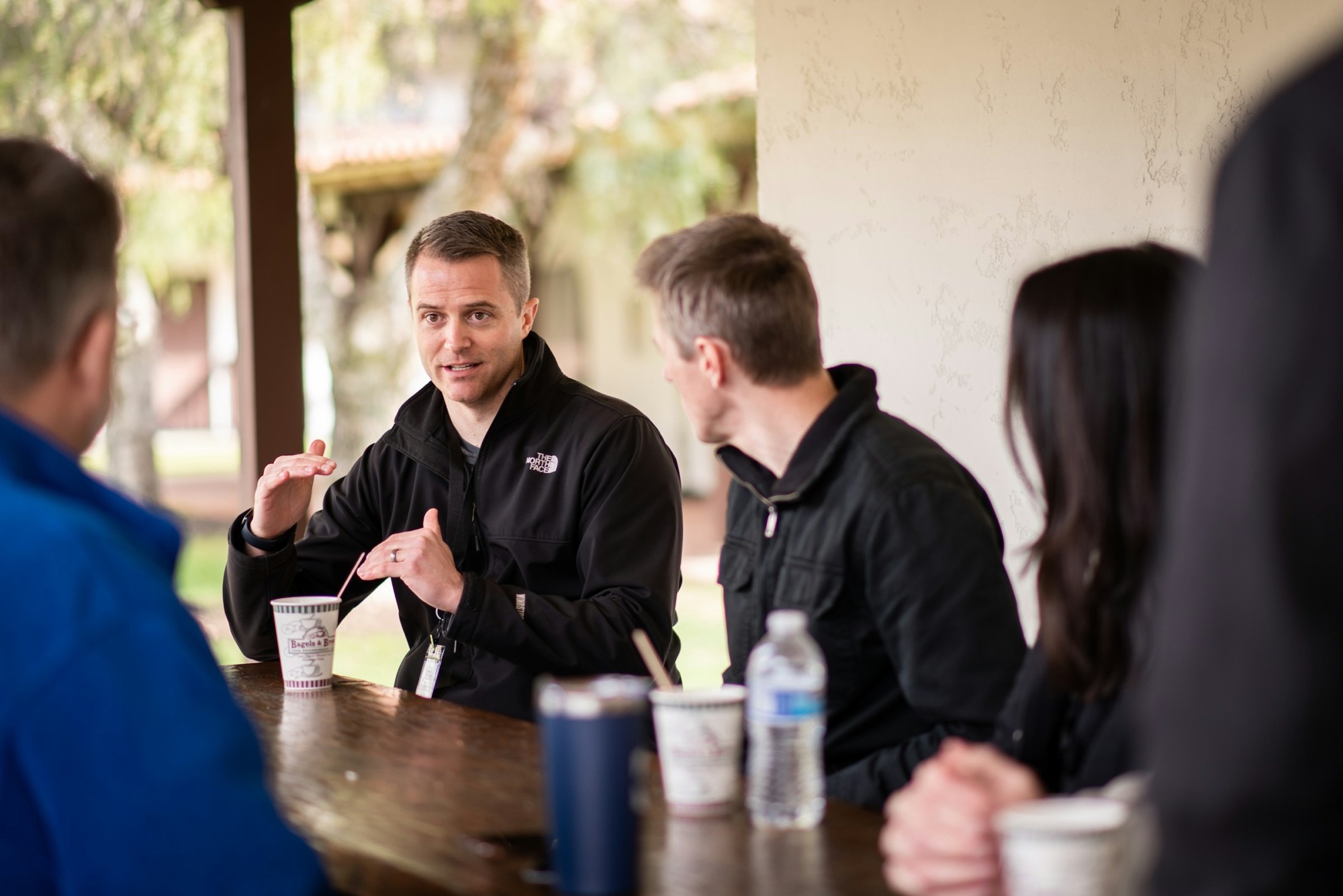 A small group talking around a wooden table outdoors under a covered porch, coffee cups and water bottles in front of them