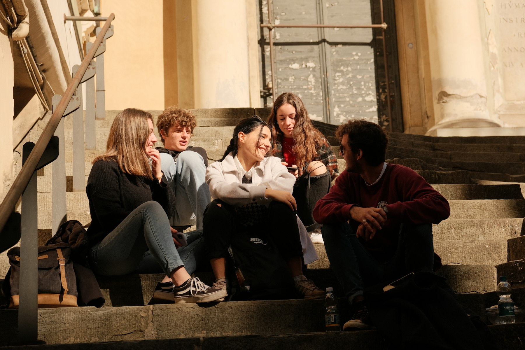 College students sitting together on sunlit stone steps outside a campus building, talking and laughing