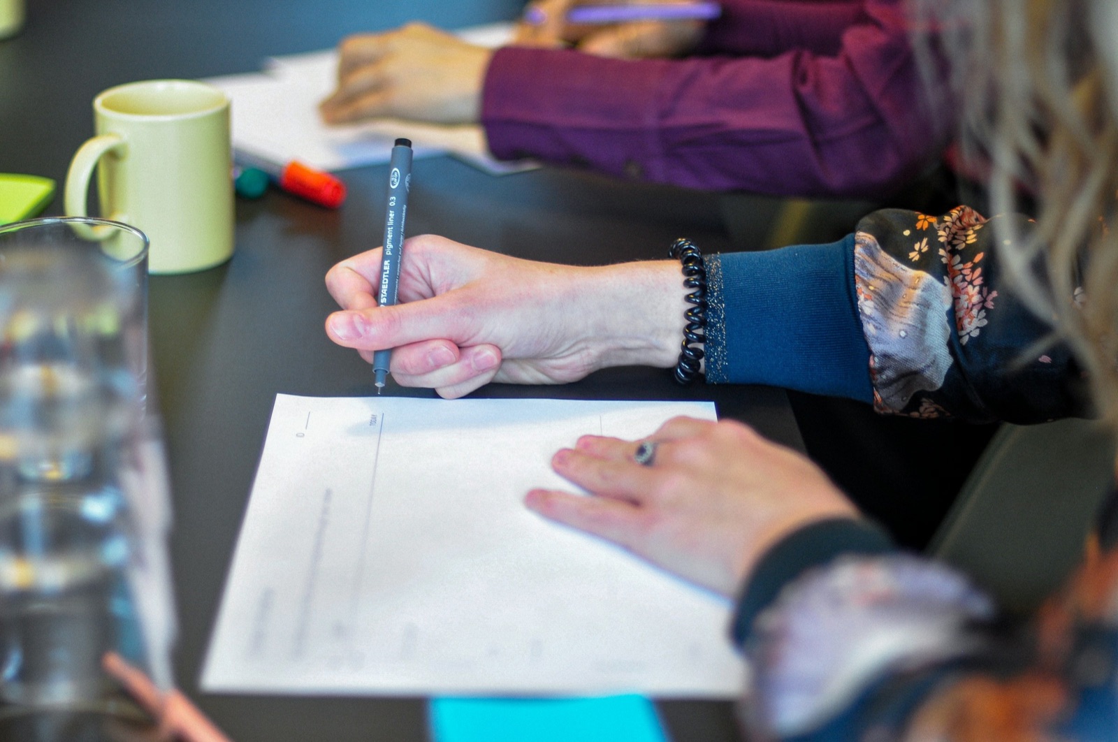 Hands writing on printed papers at a shared table, a mug and glasses nearby