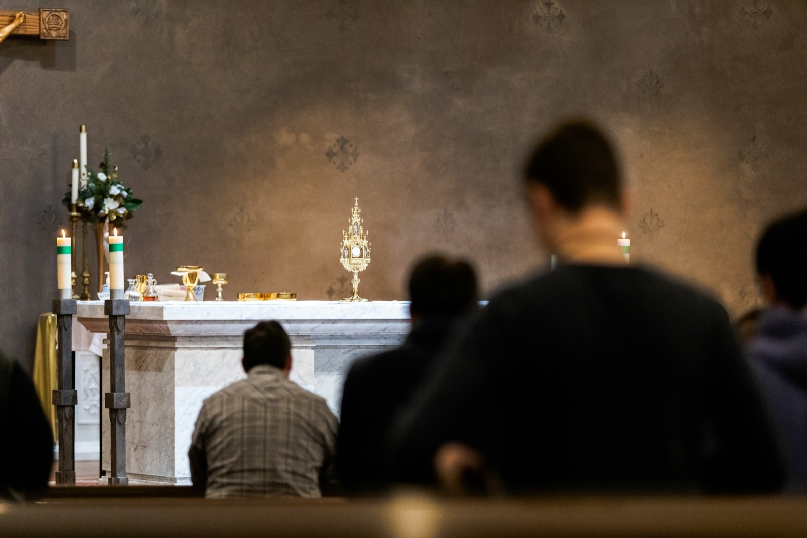 People seated in pews during a service, seen from behind, with candles and flowers at the altar
