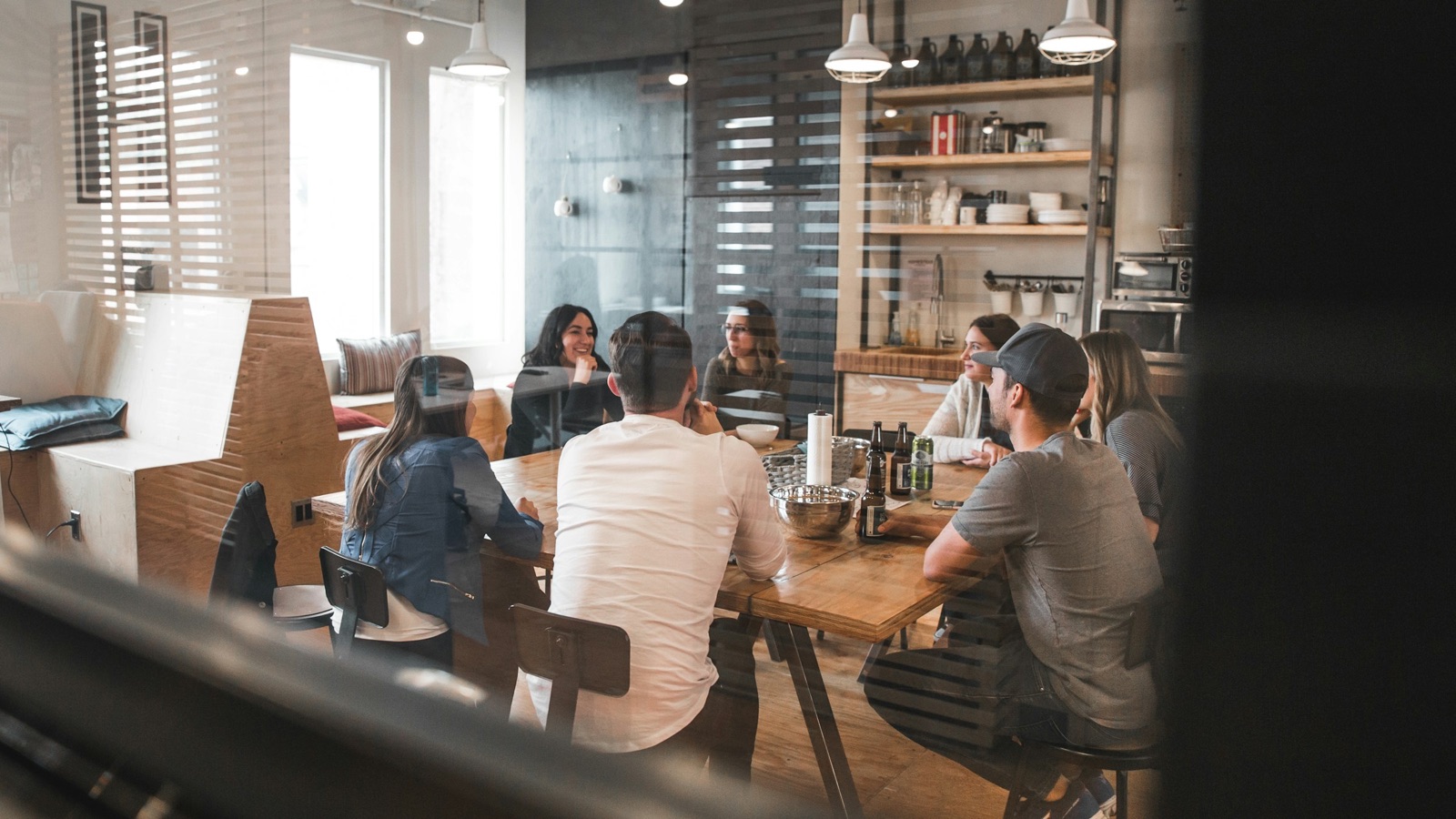 A handful of people around a long wooden table in a small studio kitchen, listening to one person talk