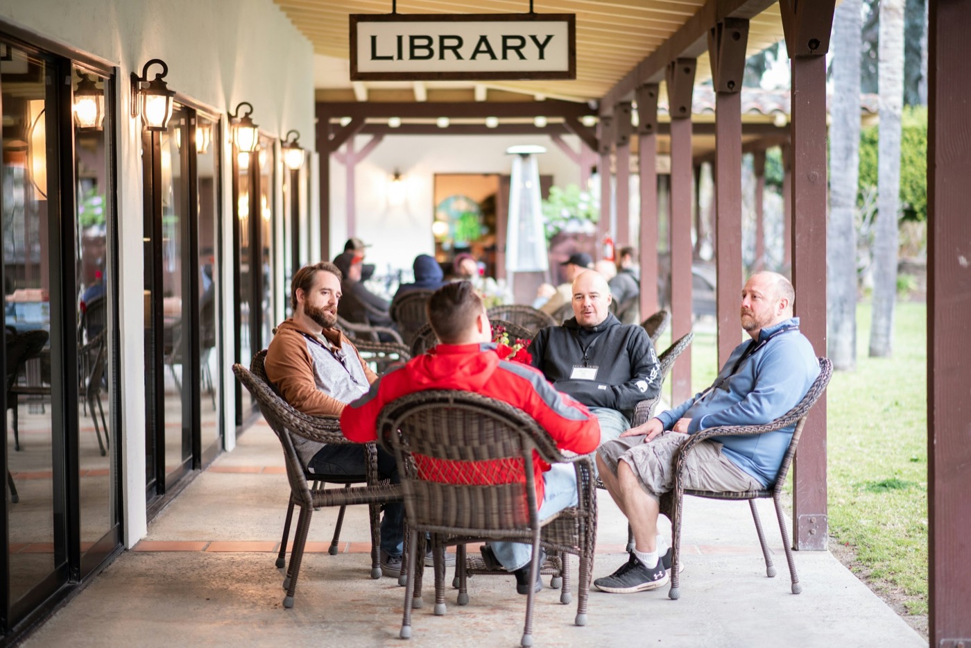 Four people talking in wicker chairs on a shaded campus porch, others gathered at tables behind them